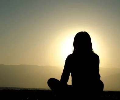Femme assise dehors, plongée dans ses pensées, entourée par la nature, visage tourné vers la lumière du jour.