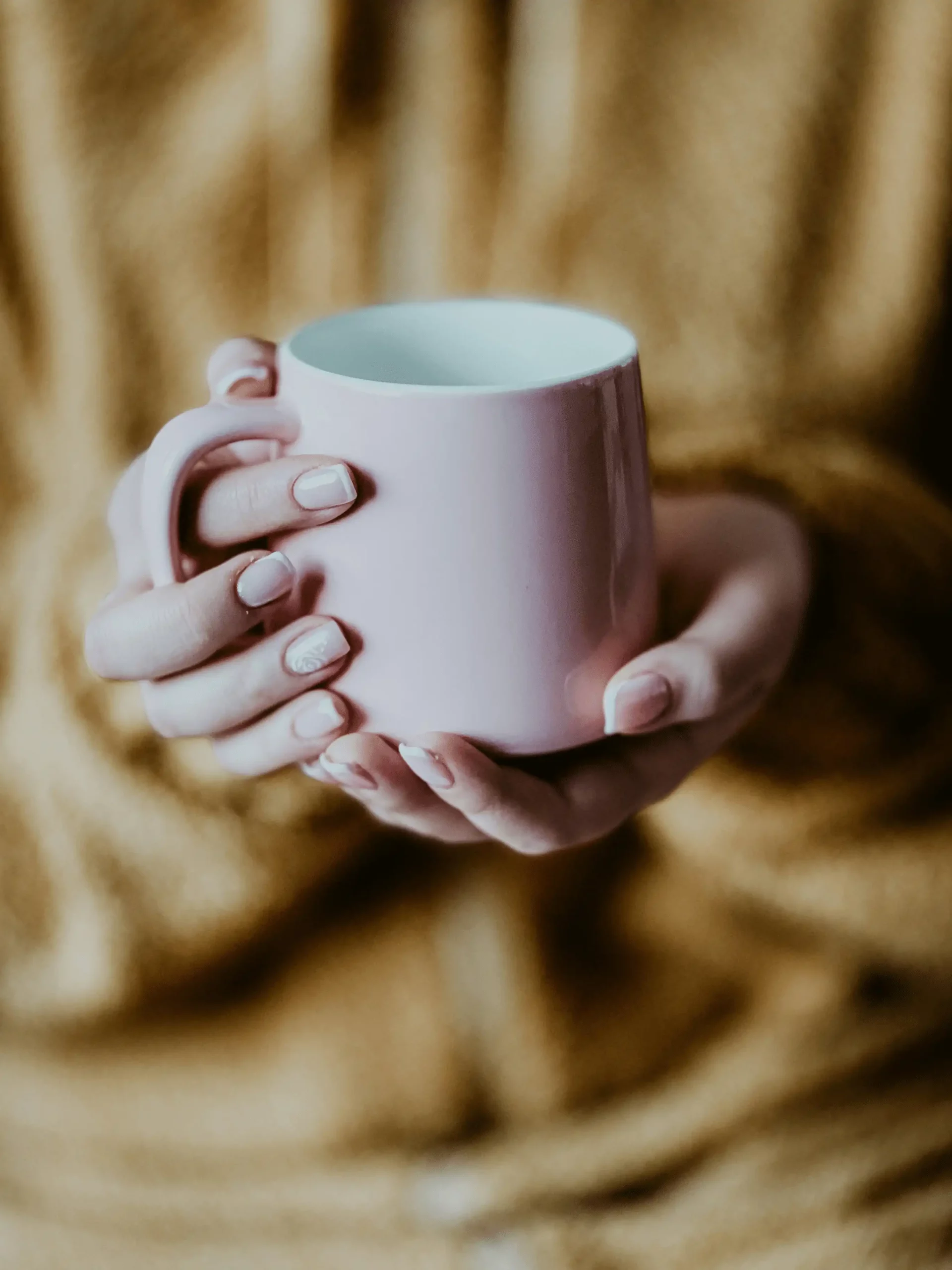 Mains de femme tenant délicatement une tasse rose en céramique dans une ambiance chaleureuse